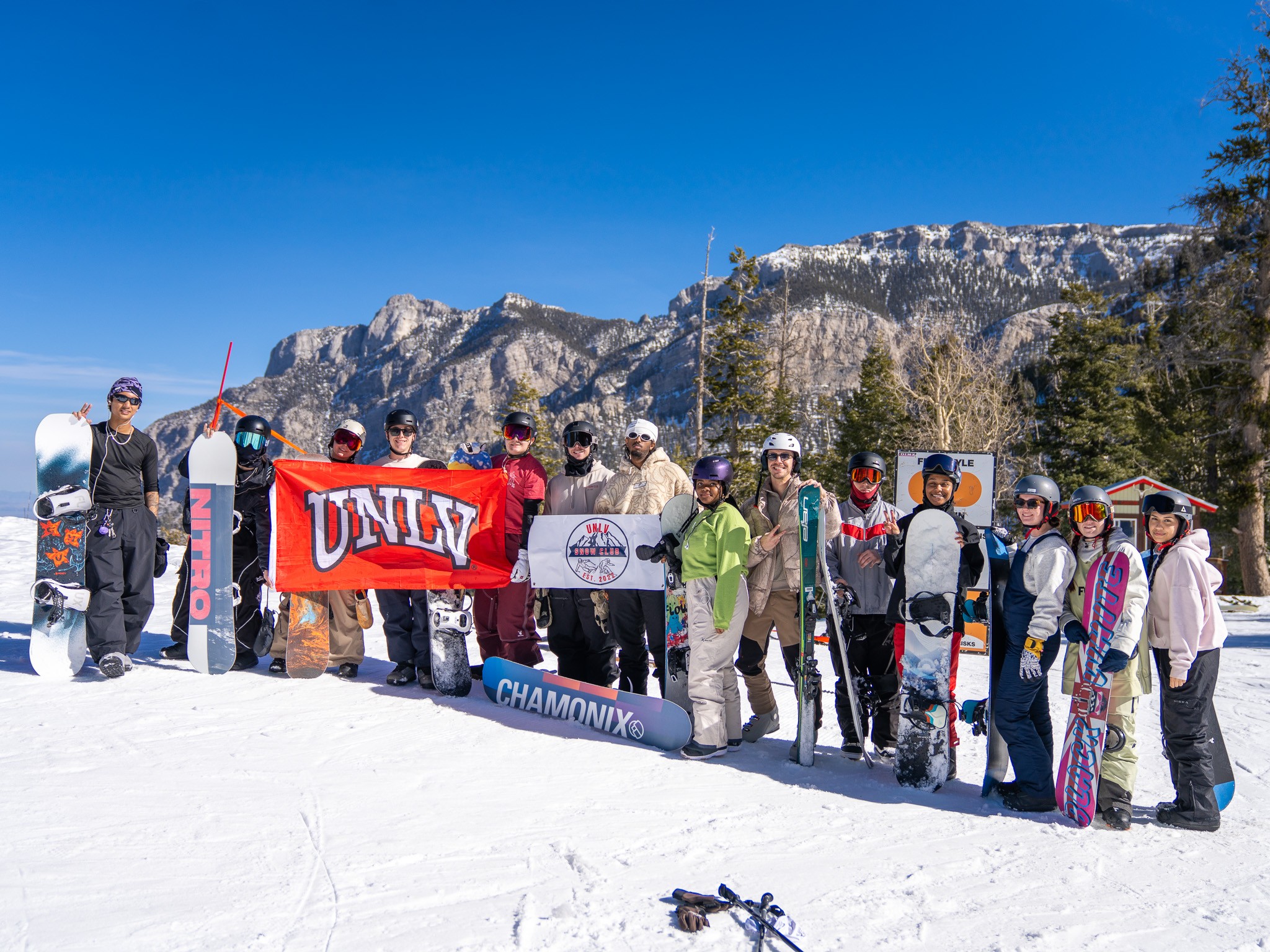 UNLV Snow Club members holding a UNLV flag and club sign on the mountain, snowboards and skis in hand